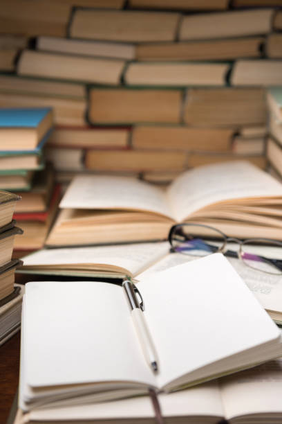 istock Education learning concept with open book and notebook on the table. Stack piles of books on reading desk and glasses in bookshelves background 1134900102