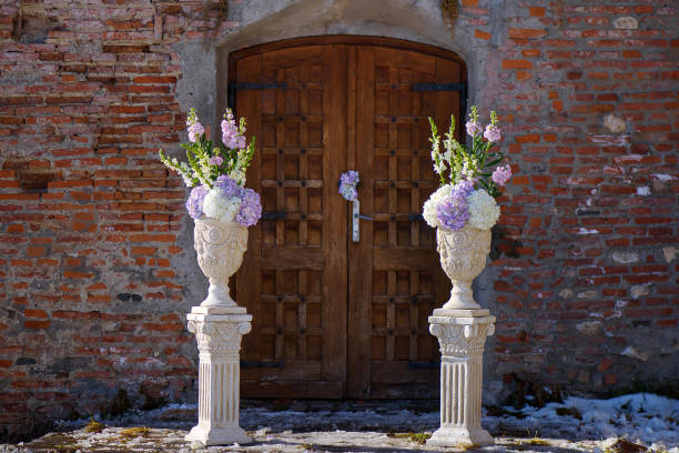 Horizontal perspective of free-standing white clay decorated flower pots with a spring bouquet featuring pastel hydrangeas positioned on each side of a vintage Medieval wooden door or gate framed by a old terracotta wall.