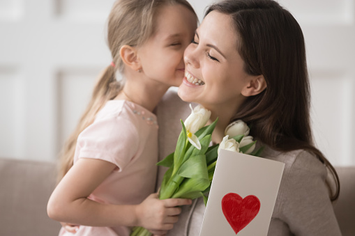 Cute little kid daughter holding spring flowers presenting greeting card congratulating happy mom with mothers day, preschool child girl embracing kiss mum make surprise to smiling mommy for birthday