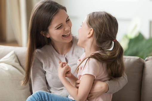 Smiling cute kid girl and loving mother having fun together on single parent day, happy family caring mom with little kid daughter playing with laughter cuddling tickling on sofa feeling joy at home