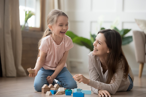 Happy family mom baby sitter and little kid daughter laughing playing with wooden blocks sit of warm floor, joyful mother having fun with child girl enjoy funny activity laughter at home together
