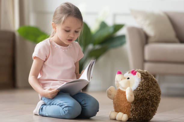 Cute little kid girl playing alone reading book to toy Cute smart little kid girl playing alone reading story to fluffy hedgehog sitting on warm floor at home, funny creative preschool small child holding book teaching toy, children imagination education girls playing stock pictures, royalty-free photos & images