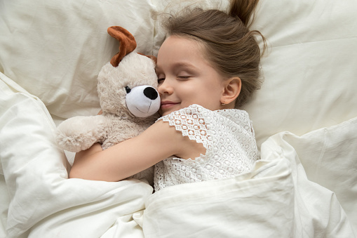 Cute little kid girl hugging teddy bear sleeping lay in cozy bed, happy small child embracing toy fall asleep on soft pillow white sheets covered with blanket having healthy night sleep, top view