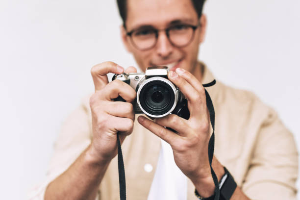 istock Image of young man smiling and taking photo on digital camera, posing over white studio background. Young male wearing casual outfit and trendy eyewear, blogging and looking to the camera display. 1134911629