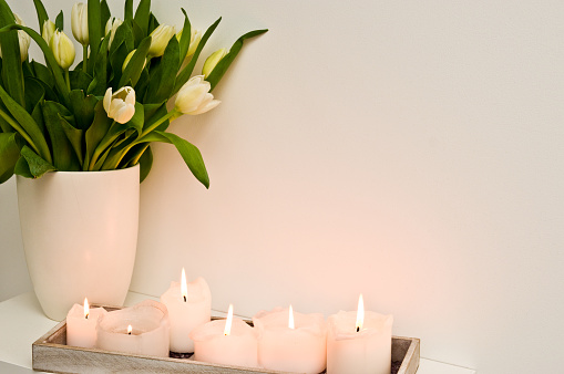 Elegant lit candles standing on a shelf against white wall, home decoration. With bouquet of white tulips.