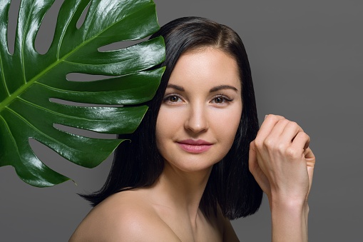 Studio beauty portrait of young brunette with natural make-up perfect skin with green exotic leaf on gray background, copy space. Concept of natural cosmetics, face and body care products