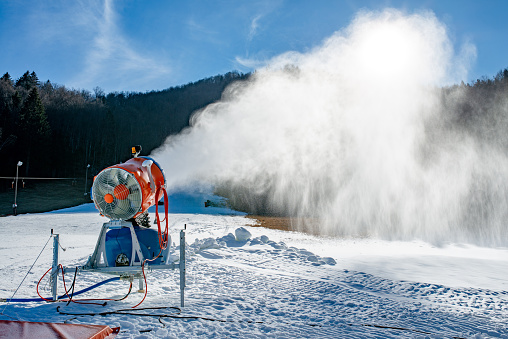 Snow machine blowing artificial snow over a ski slope on a sunny day