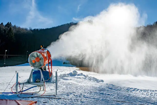 Photo of Snow machine blowing artificial snow over a ski slope on a sunny day