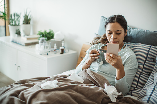 Woman with common cold in bed, she use home medicine to handle sickness. She using mobile phone