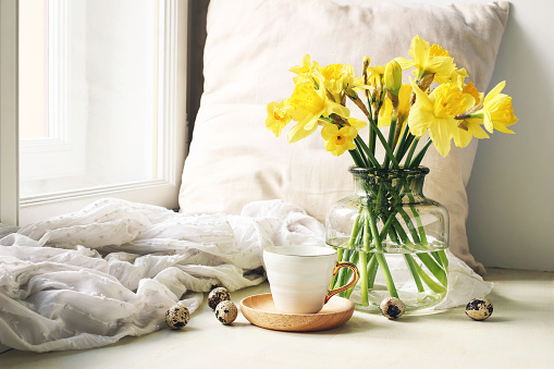 Cozy Easter, spring still life scene. Cup of coffee, wooden plate, quail eggs and vase of flowers on windowsill. Floral composition with yellow daffodils, narcissus. Vintage feminine styled photo.