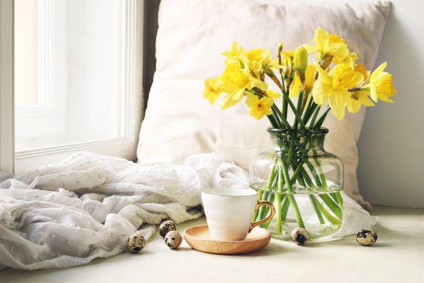 cozy easter, spring still life scene. cup of coffee, wooden plate, quail eggs and vase of flowers on windowsill. floral composition with yellow daffodils, narcissus. vintage feminine styled photo. - daffodil flower yellow vase imagens e fotografias de stock