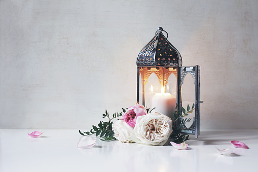 Vintage silver Moroccan, Arabic lantern with glowing candle, green branches, rose flowers and pink petals on white table background, greeting card for Muslim holiday Ramadan Kareem. Shaby wall.