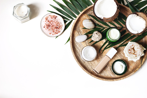 Styled beauty composition. Skin creams, makeup bottle, rose and pebble stones on wooden tray. Coconuts, tropical palm leaves decoration. Cosmetics, spa concept, empty space, flat lay, top view.