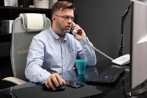 representative man with facial hair in gray shirt and jeans works in modern office at computer. businessman answers call on landline. Portrait of entrepreneur at work in office