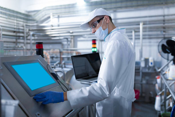 mann in einer weißen robe und eine maske mit einem laptop in den händen steht in der nähe des digitalen bildschirms in der fabrik - lebensmittelverarbeitung stock-fotos und bilder