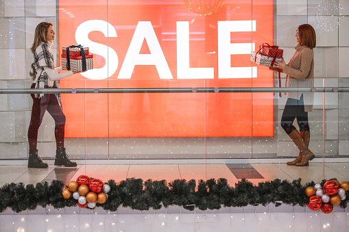 Side view of a mature woman and her daughter standing by the large SALE sign in shop window, holding gift boxes.