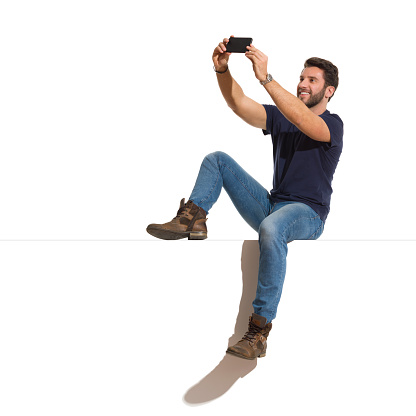 Young man in boots, jeans and unbuttoned lumberjack shirt is sitting on a top, holding telephone and taking a selfie. Full length studio shot isolated on white.