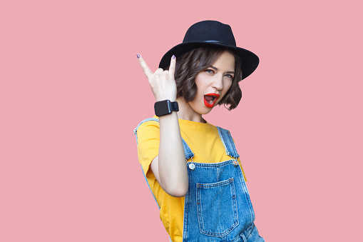 Portrait of beautiful amazed young woman in yellow t-shirt, blue denim overalls with makeup and black hat standing with rock horns, screaming and looking at camera. studio shot on pink background.