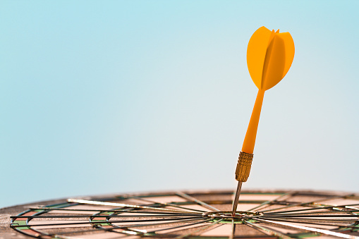 Orange dart arrow hitting bullseye in center target of dartboard on blue sky background with copy space. Business, strategy, achievement, and planning concept