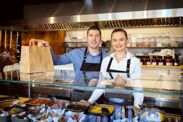 Photo of Portrait of small business owner smiling behind counter inside eatery, Successful young waiters working in apron holding food order for home delivery