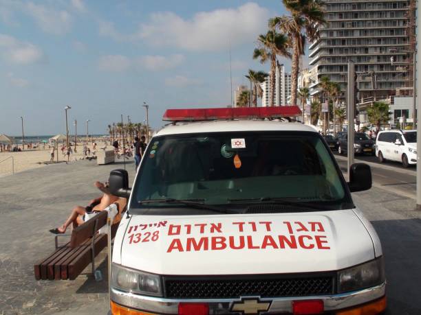 Israeli Ambulance Plus People View At Beach Promenade In Tel Aviv. Israel View Of Packed Israeli Ambulance, Building Exterior, Vehicle On The Road, People Playing And Swimming On The Beach, Walking Around, Sitting Down On Beach Promenade During The Day In Tel Aviv Israel ambulance in israel stock pictures, royalty-free photos & images