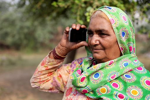 Senior women of Indian ethnicity standing outdoor in nature & talking on mobile phone.