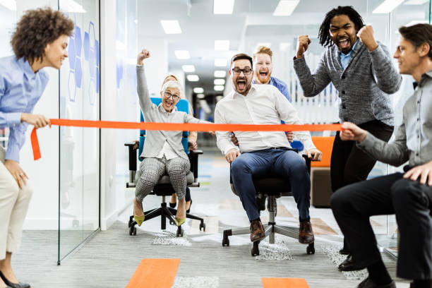 Large group of joyful colleagues having fun during chair race in the office. Cheerful business team having fun during chair race in the office. office competition stock pictures, royalty-free photos & images