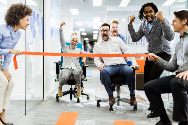 Large group of joyful colleagues having fun during chair race in the office. Cheerful business team having fun during chair race in the office. race office stock pictures, royalty-free photos & images