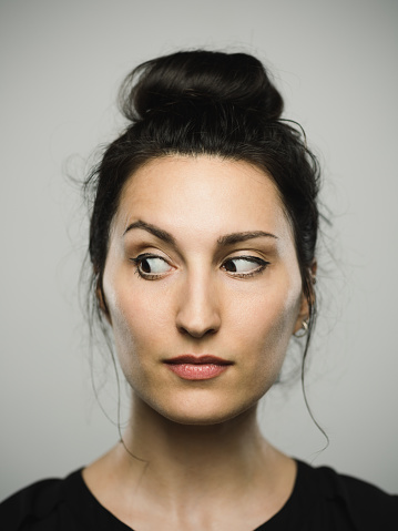 Close up portrait of mediterranean young woman looking to the side against gray white background. Vertical shot of caucasian real people observing in studio with long black hair and a hair bun. Photography from a DSLR camera. Sharp focus on eyes.