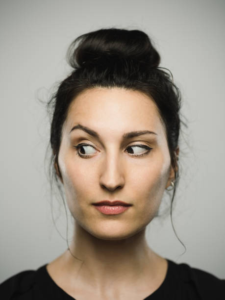 Studio portrait of real mediterranean young woman looking to the side Close up portrait of mediterranean young woman looking to the side against gray white background. Vertical shot of caucasian real people observing in studio with long black hair and a hair bun. Photography from a DSLR camera. Sharp focus on eyes. suspicion stock pictures, royalty-free photos & images