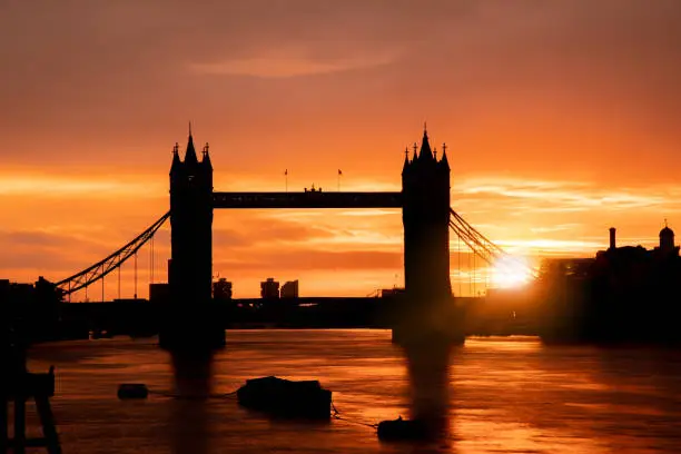 Tower Bridge & Thames river in city of London at dawn. Backlit contrajour silhouette photo of the iconic architecture of the famous suspending bridge, photographed from the river Thames North shore to compose the rising sun flare. Shot on Canon EOS R full frame system. Image is ideal for background with plenty of copy space.