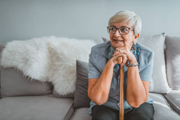 No more balancing scares for me! Photo of Retired short hair old woman with her walking stick at home looking away during the day while sitting on sofa. Senior gray hair women supporting on a walking cane at home. walking stick stock pictures, royalty-free photos & images