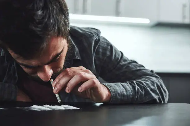Photo of Drug addiction is a self-punishing disease that ruins mental, psychological and physical health. Man snorts a line of cocaine with a rolled up bank note. Close up