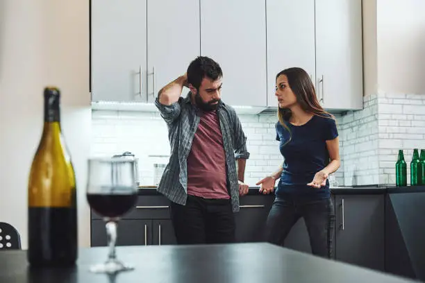 Portrait of man and woman having argument, standing in the kitchen. A bottle and a glass of wine are on the table. Selective focus