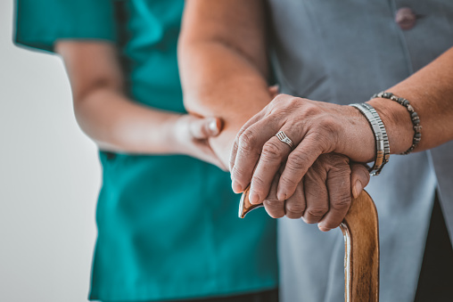 Photo of Unrecognizable Senior Woman's Hands On Walking Stick With Care Worker In Background. Young woman holding hand of old woman with walking stick. Female nurse supporting senior woman in with walking stick at home during the day.