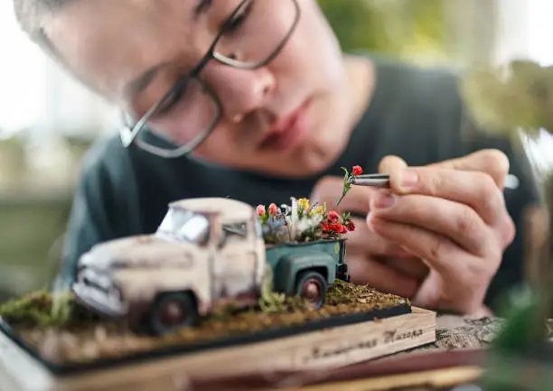 The young man have a hobby to make a miniature dioramas and a scale models. A young artist is sitting at the table in his home workshop. He is decorating with the small rose the retro pickup truck. Close-up shooting