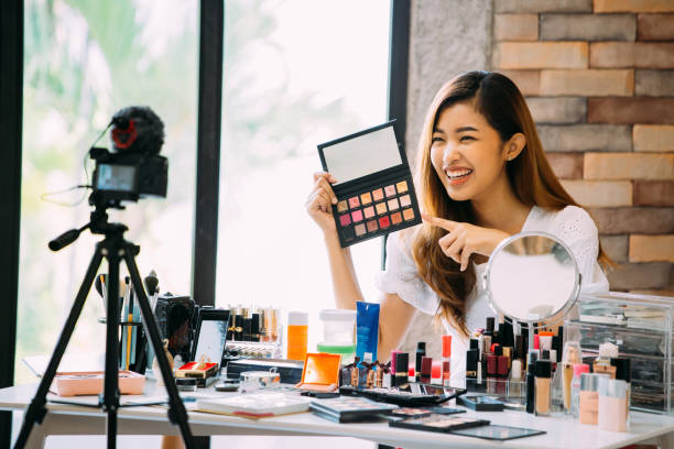Charming Asian girl recording vlog. Beauty blogger presenting makeup cosmetics in front of camera Pretty Asian woman sitting at table and making video about cosmetics while filming with camera on tripod tutorial photos stock pictures, royalty-free photos & images