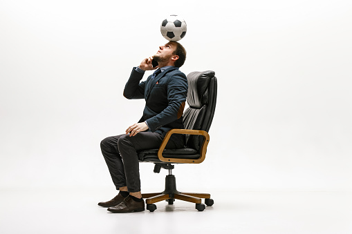 Businessman with football ball in office. Soccer freestyle. Concept of balance and agility in business. Manager perfoming tricks while sitting on chair and speaking on the smartphone isolated on white studio background.