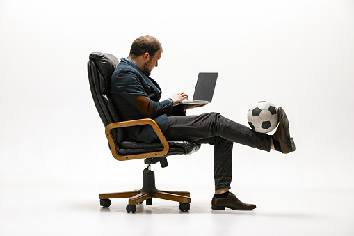 Businessman with football ball in office. Soccer freestyle. Concept of balance and agility in business. Manager perfoming tricks sitting on chair and working on laptop isolated on white studio background.