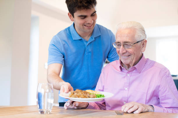 asistente de cuidado masculino sirviendo comida a senior masculino sentado en la mesa - atención residencial fotografías e imágenes de stock