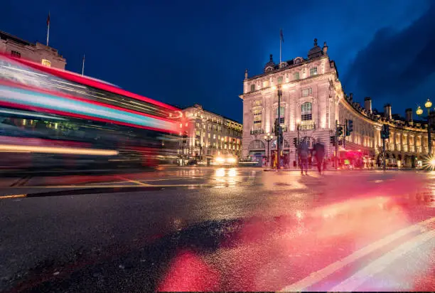 Long exposure photo with evening illumination at Piccadilly Circus in London city during blue hour on a rainy English evening. Crowd and traffic with motion blur due to the exposure time with unrecognisable accidental commuters and tourist. Image has copy space and it is ideal for background. Shot on Canon EOS R full frame system.