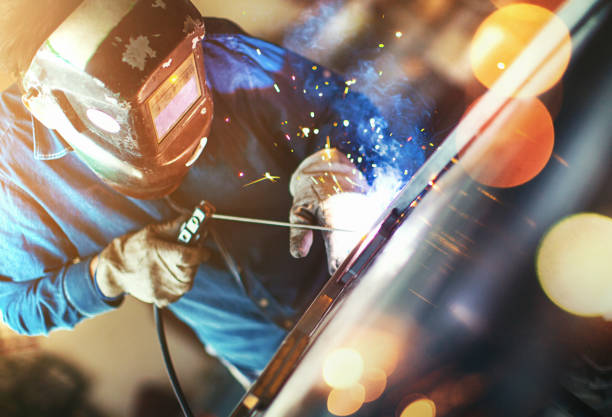 Welding process. Closeup side view of unrecognizable worker welding two metal plates with gas welding machine.He's wearing welding mask and protective workwear and gloves. Sunlight is coming from the right. welding helmet stock pictures, royalty-free photos & images