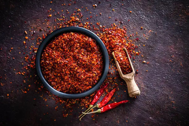 Spices: Top view of a black bowl filled with red chili pepper flakes shot on abstract brown rustic table. A wooden serving scoop with pepper flakes is beside the bowl, pepper flakes are scattered on the table. Three dried red chili peppers are beside the bowl. Predominant colors are brown and red. Low key DSRL studio photo taken with Canon EOS 5D Mk II and Canon EF 100mm f/2.8L Macro IS USM.