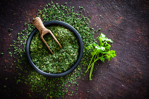 Spices: Top view of a black bowl filled with dried parsley shot on abstract brown rustic table. A wooden serving scoop is inside the bowl and dried parsley is scattered on the table. A small bunch of fresh organic parsley is beside the bowl. Predominant colors are brown and green. Low key DSRL studio photo taken with Canon EOS 5D Mk II and Canon EF 100mm f/2.8L Macro IS USM.