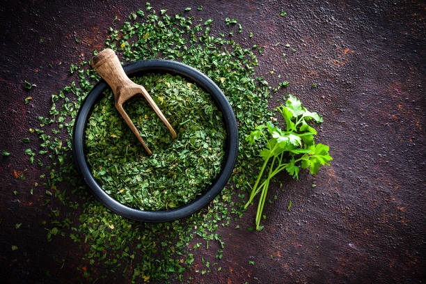 Dried parsley shot from above Spices: Top view of a black bowl filled with dried parsley shot on abstract brown rustic table. A wooden serving scoop is inside the bowl and dried parsley is scattered on the table. A small bunch of fresh organic parsley is beside the bowl. Predominant colors are brown and green. Low key DSRL studio photo taken with Canon EOS 5D Mk II and Canon EF 100mm f/2.8L Macro IS USM. parsley stock pictures, royalty-free photos & images