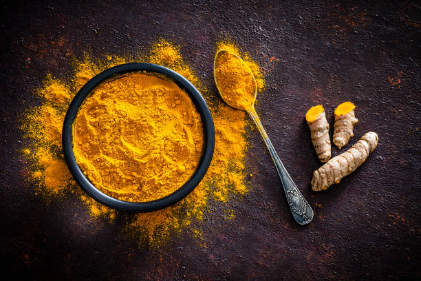 Spices: Turmeric roots and powder shot from above Spices: Top view of a black bowl filled with turmeric powder shot on abstract brown rustic table. A metal spoon with turmeric powder is beside the bowl and turmeric powder is scattered on the table. Fresh organic turmeric roots are beside the spoon. Predominant colors are brown and yellow. Low key DSRL studio photo taken with Canon EOS 5D Mk II and Canon EF 100mm f/2.8L Macro IS USM. curry stock pictures, royalty-free photos & images