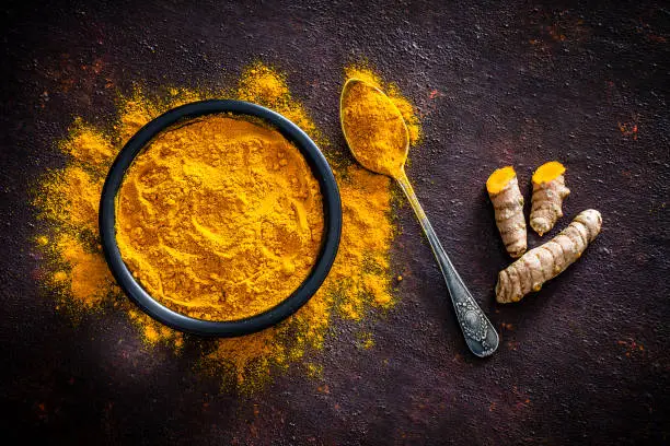 Spices: Top view of a black bowl filled with turmeric powder shot on abstract brown rustic table. A metal spoon with turmeric powder is beside the bowl and turmeric powder is scattered on the table. Fresh organic turmeric roots are beside the spoon. Predominant colors are brown and yellow. Low key DSRL studio photo taken with Canon EOS 5D Mk II and Canon EF 100mm f/2.8L Macro IS USM.