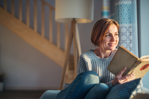 A cheerful young woman sitting indoors on a sofa at home, reading a book. Copy space.