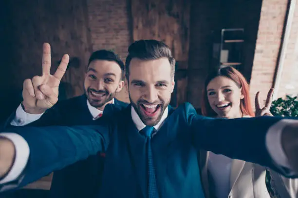 Photo of Self-portrait of three nice stylish beautiful handsome cheerful crazy optimistic executive managers showing v-sign enjoy great day pause break in loft industrial interior work place station