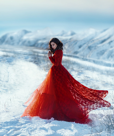 The girl in a luxurious, lush, dress with a train of red, walks against the backdrop of a winter mountain landscape. Emotions of sadness and cold. Lonely brunette woman with a middle-aged novel